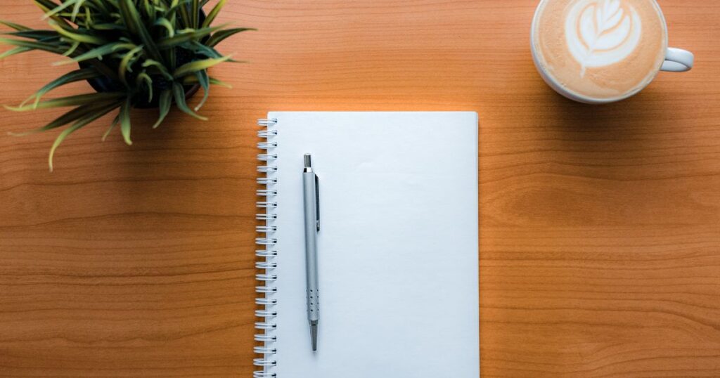 Carnet à spirale vierge et stylo sur un bureau en bois, à côté d’une tasse de café et d’une plante